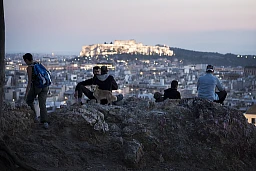 Junge Leute sitzten im Quartier Exarchia auf einem Hügel und blicken südwesten über Athen und die Akropolis und Parthenon im Hintergrund.